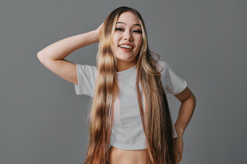 Young woman with long hair and white crop top smiling and posing with hand behind head on gray...