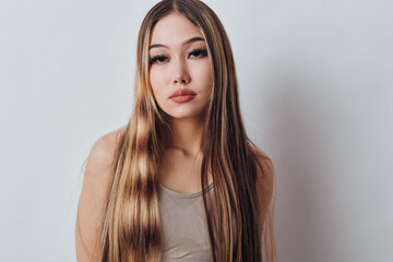Fototapeta premium Young woman with long straight hair posing against white background with natural makeup and neutral expression wearing sleeveless top indoors.
