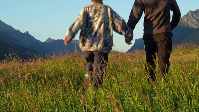Two people walk hand in hand through a lush green mountain meadow at sunrise, with clear skies. Waterton, AB, Canada