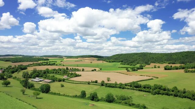 Aerial view of the French countryside with patchwork fields, green trees, and scattered farm buildings under a bright blue sky with fluffy clouds.