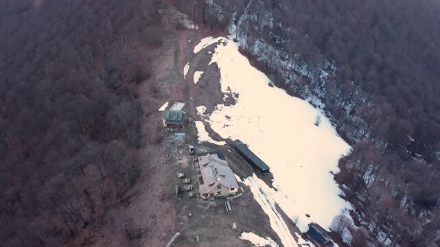 Aerial drone tilt up from Mazalat Hut to surrounding mountain peaks in Stara Planina, Bulgaria. Early spring landscape with patchy snow and forested ridges under soft evening light.