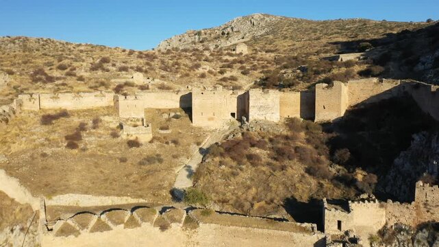 Wide view of the historic Acrocorinth fortress ruins set on a rugged, sunlit hillside in Greece. The scene features stone walls and arid Mediterranean landscape under a clear blue sky.