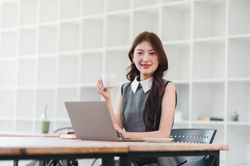 Happy young asian woman business professional sitting at desk with laptop holding coffee cup in...