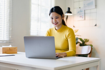 Fototapeta premium Cheerful young asian woman wearing yellow sweater working with laptop computer on white desk in bright home office with happy smile