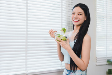 Happy young Asian woman eating fresh healthy salad vegetable near window blind. Smiling female enjoys nutrition diet lifestyle with organic food