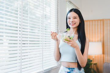 Happy young asian woman eating fresh healthy green salad vegetable from bowl at home near window blind. Female smile with wellness diet lifestyle