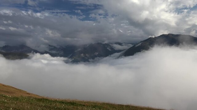 Mountain valley blanketed by low clouds above alpine meadow, dramatic sky and peaks. Misty ridge scene with overcast weather, rolling fog layer, remote highlands, rugged summits.