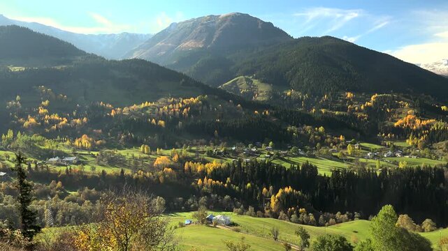 Alps village in mountain valley with autumn forest, green meadows, panoramic cinematic landscape. Scenic hamlet among fall woods and rolling highlands, fairy tale countryside vista, vivid.