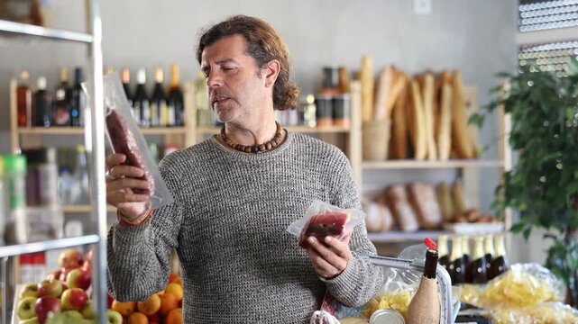Attentive and interested man with basket full of groceries examines salami and meat in sausage department of small supermarket