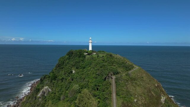 Farol das Conchas lighthouse on Ilha do Mel near Paranagu&aacute;, Paran&aacute;, Brazil, standing on a forested hill above the Atlantic Ocean coastline with visible ridge trail leading to the tower, drone push in