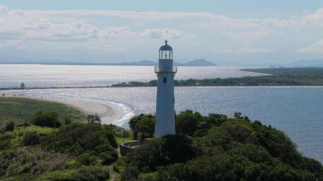 White Faro das Conchas lighthouse Farol das Conchas stands on Ilha do Mel near Paranagu&aacute;, Paran&aacute;, Brazil, surrounded by Atlantic coastal scrub with beach and bay waters behind, drone orbit shot.
