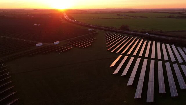 Aerial drone sequence of a sustainable solar power facility in England at sunset featuring dramatic red light reflections on the panel rows.