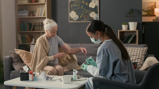 Brunette health worker dressed in uniform drawing up fluid from vial into disposable syringe while elderly lady exposing her shoulder, then girl cleansing injection site