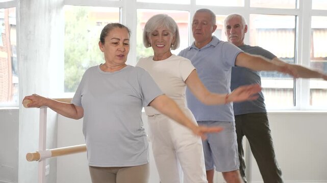 Positive old female attendee of ballet courses doing battement tendu standing by training barre with others