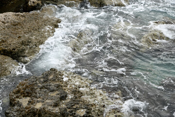 High-angle shot of ocean waves crashing against rugged coastal rocks. The churning seawater creates white foam and spray over the dark, weathered stone surface of a Mediterranean shoreline.