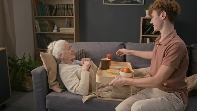 Caring teenage grandson bringing portable bed tray with tea mug, medications and citrus fruits for sick granny lying on couch