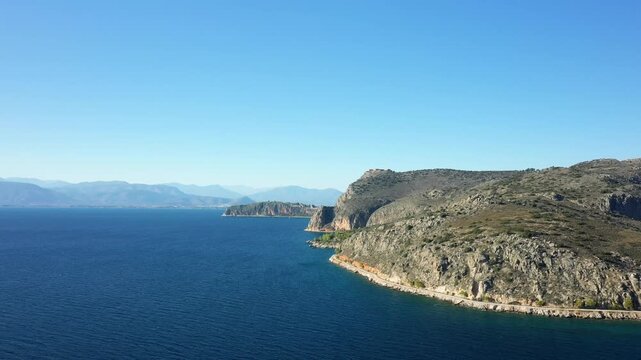 Scenic view of the rocky coastline and deep blue sea under a clear sky in Nafplio, Greece. Rugged cliffs meet the tranquil waters, creating a serene Mediterranean landscape.