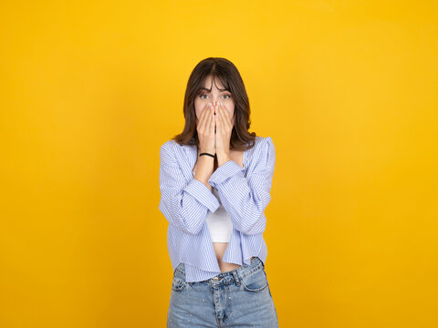 Shocked young woman covering her mouth with hands in amazement, looking at camera with wide eyes against yellow background. Concept of secret, surprise, disbelief, and unexpected news.
