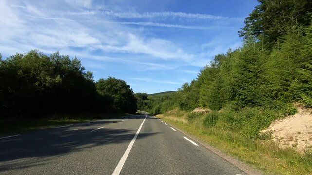 Empty country road winding through lush green forest in the Morvan region of France under a blue sky with wispy clouds. Peaceful rural landscape ideal for travel and nature concepts.
