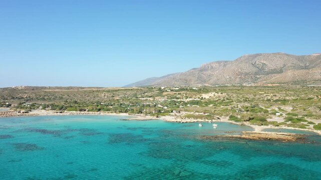 Crystal clear turquoise waters and sandy shores of Elafonisi beach in Crete, Greece, with rugged mountains in the background under a clear blue sky.