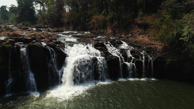 Beautiful cascading Tad Lo waterfall surrounded by lush forest and rocky terrain in southern Laos. Tranquil natural landscape with flowing water and greenery.