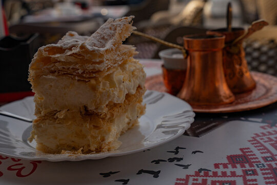 Epic Bosnian krempita cake and coffee authentic texture. 
Cinematic close up of traditional cream cremschnitte pastry, 
copper cezve and national tablecloth in old town cafe.
