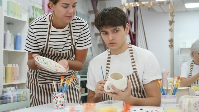 Cheerful young couple, guy and girl, actively engaged in creative hobby, decorating ceramic pottery together at table in cozy workshop setting