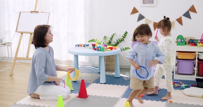 Asian female teacher kneel on mat, hold ring toss set, Asian preschool girl reach for ring while Asian preschool boy run past, colorful cone marker lane, active daycare play, coaching motor skill.