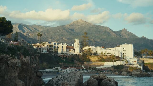 Golden hour view of Nerja tourist city with whitewashed buildings, church tower, rocky cliffs and Mediterranean Sea framed by dramatic Andalusian mountains.