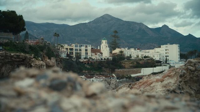 Scenic view of Nerja along the southern coast of Spain, framed by rocky foreground elements with white buildings and a church tower set against dramatic Andalusian mountains.