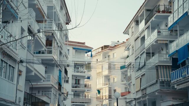 White residential apartment buildings with balconies, blue awnings, and visible overhead utility wires in a Mediterranean coastal town.