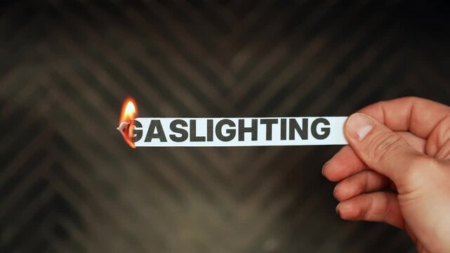 Hand holds a paper strip reading &ldquo;GASLIGHTING&rdquo; as the edge burns in flame, symbolizing psychological manipulation, toxic relationships, and emotional abuse. Concept b-roll.