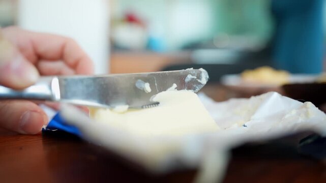 Close-up hand scoops butter from an opened wrapper with a knife on a wooden table, preparing ingredients for breakfast toast. Natural light food b-roll.