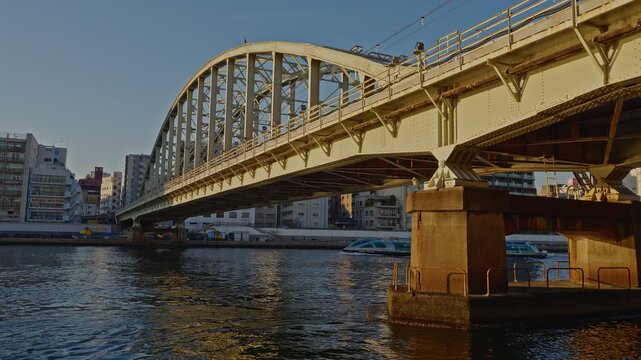A wide shot of the JR Sobu Line Sumidagawa Railway Bridge, a historic steel arch bridge in Tokyo, with a water bus passing underneath on the Sumida River at sunset.
