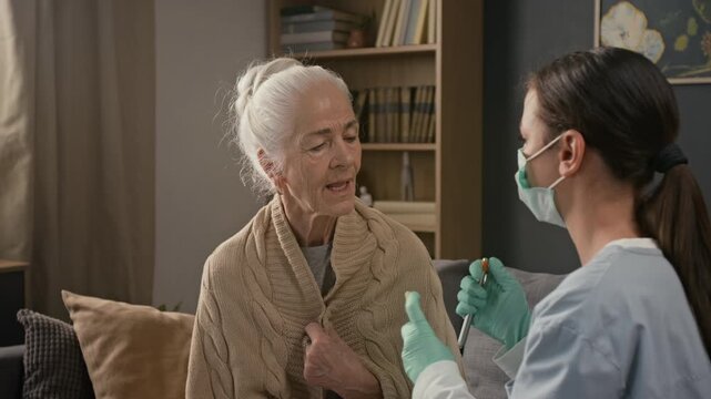 Young health worker wearing face mask using spatula and diagnostic penlight to examine throat of elderly woman during home care visit