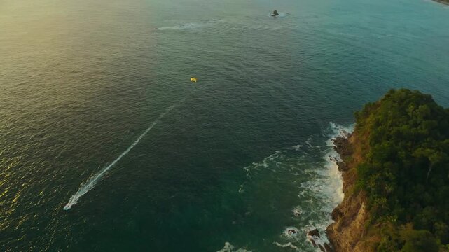 Drone view of a parasailing boat gliding along cliffs over turquoise ocean waters, bathed in warm sunset glow