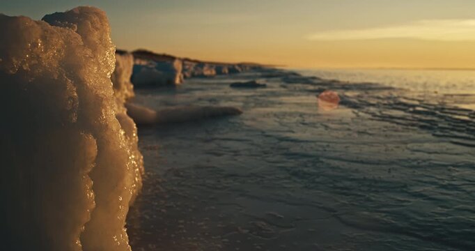 Icy winter beach at the Curonian Spit, Lithuania, as Baltic Sea waves drift inshore at golden hour. Frozen shoreline textures, warm sunset light, and calm cold coastal atmosphere