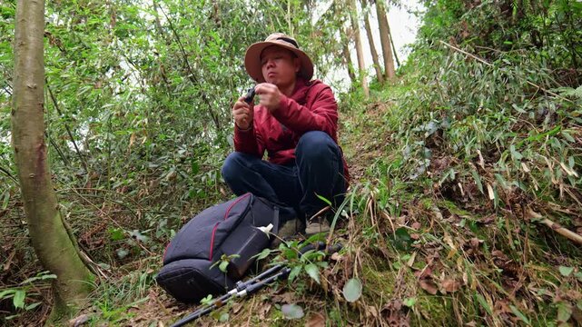 A man hiking in the uninhabited mountain forest squats on the ground to eat chocolate for energy. A man hiking in the primeval forest is having a picnic.