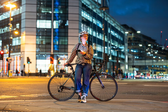 Woman with bicycle using phone at city crosswalk in London at night