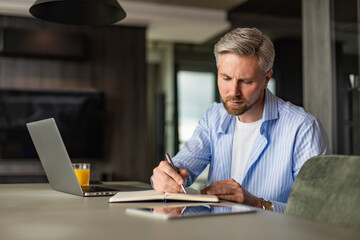 Fototapeta premium Young businessman sitting at home working on a laptop and writing notes down on a notepad.