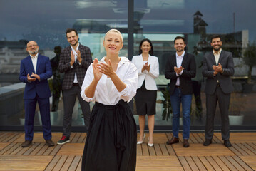 Fototapeta premium Smiling businesswoman stands on a rooftop terrace as colleagues and a diverse business team applaud her leadership in the corporate office. Business people group celebrating success. 