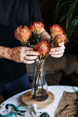 Person holds orange pincushion protea flower while arranging bouquet in glass vase on table. art therapy, tactile mindfulness, sensory wellness, therapeutic gardening