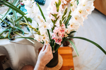 Male hand adjusts white and pink gladiolus flowers in dark glass vase. Flower arranging for anxiety, tactile wellness, plant-based healing, emotional well-being.