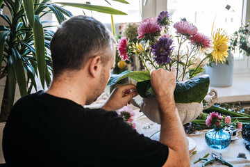 Man arranges vibrant asters in vintage white urn on studio table. Stress management for men, therapeutic floristry, hobby-based mental health, cortisol reduction, mindful craftsmanship.