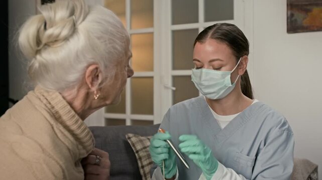 Positive nurse wearing face mask, uniform and rubber gloves showing spatula to senior lady and checking her throat using medical penlight while providing home care services