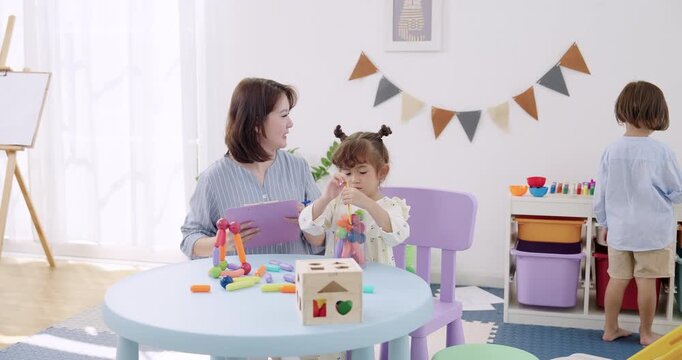 Asian female preschool teacher reviewing clipboard notes while toddler boy steps away from table, toddler girl stands near toys, daycare routine, child care supervision, learning progress tracking.