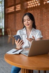 Fototapeta premium Pretty asian woman holding and looking at phone over book and laptop sitting at wooden table in cafe