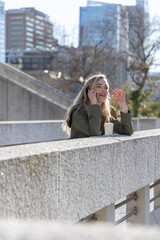 Woman in 20s leaning on railing at terrace, talking on smartphone, olive green jacket, takeaway cup