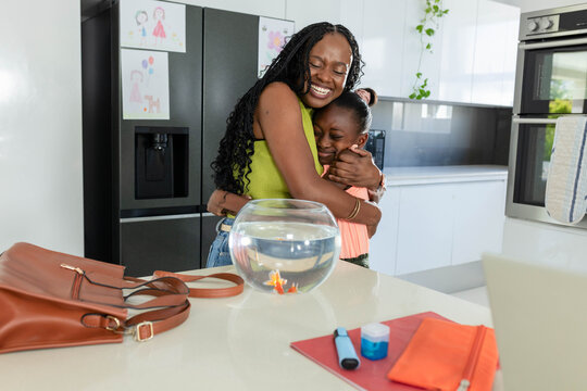 African mother in lime green top hugging two youth daughters in modern kitchen near fishbowl