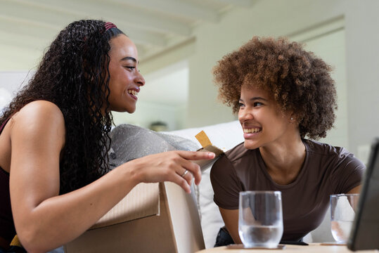 African American female friends sitting on sofa at home, leaning on moving box, sharing tea tag